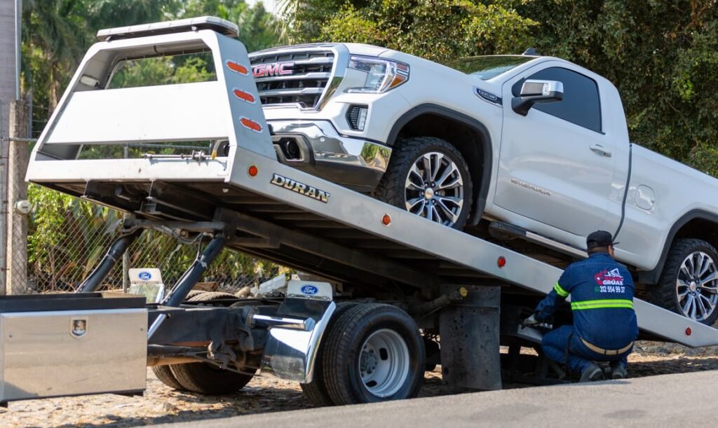Tow truck operator loading white GMC pickup truck on street in daytime.